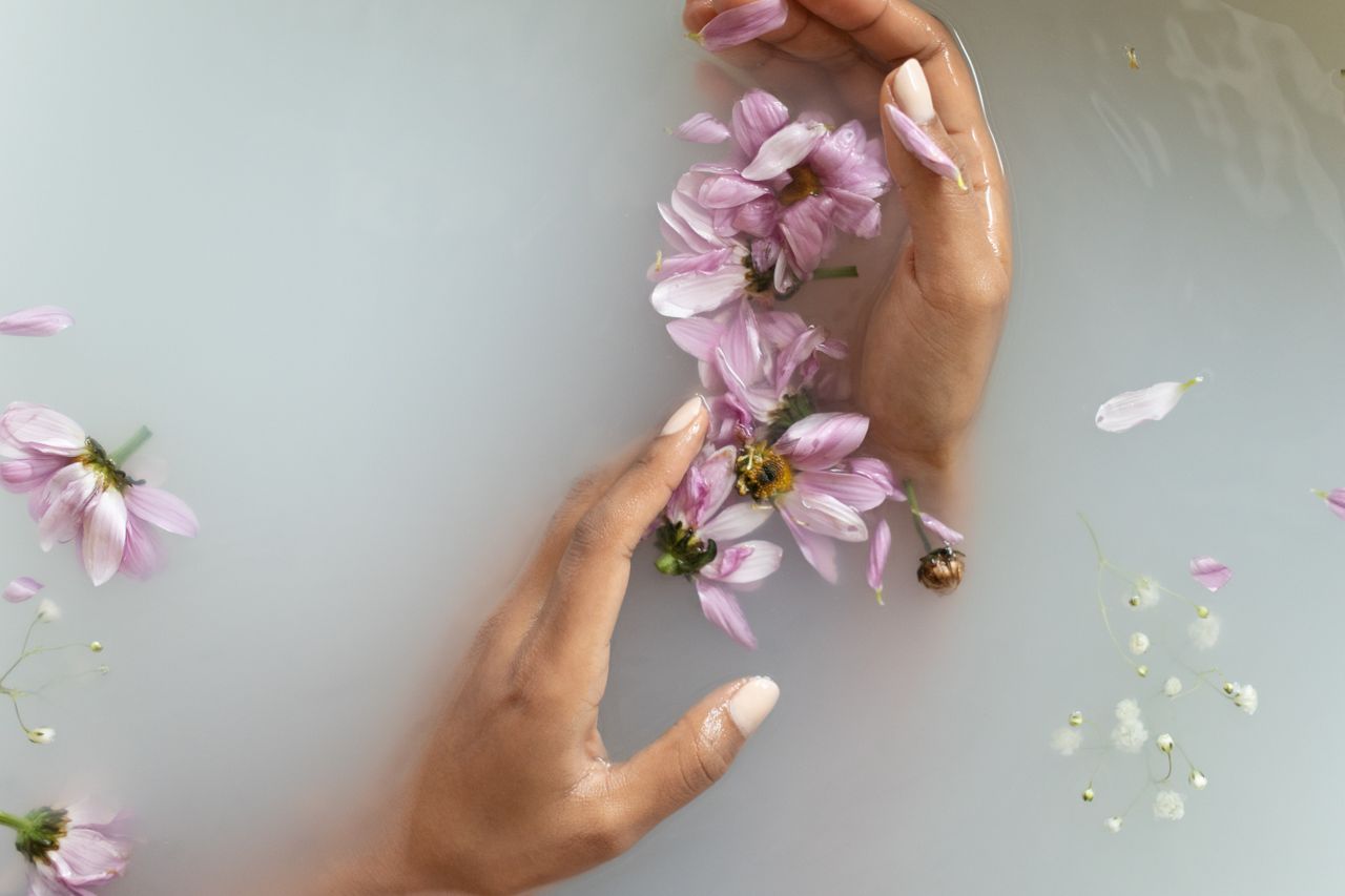 Home Top view of anonymous female with manicured hands touching delicate flowers while taking bath with white soapy water